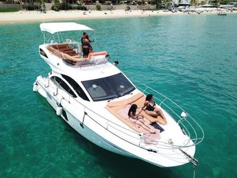 White luxury yacht anchored in clear turquoise water off a sandy beach, with two people sunbathing on the bow and others on the upper deck enjoying a sunny coastal vacation.