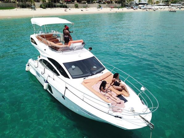 White luxury yacht anchored in clear turquoise water off a sandy beach, with two people sunbathing on the bow and others on the upper deck enjoying a sunny coastal vacation.
