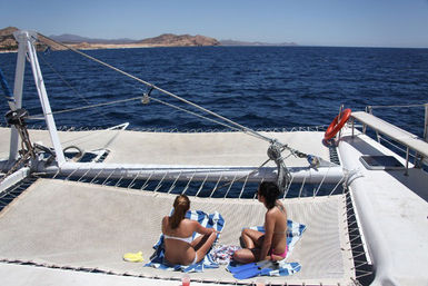 Two sunbathers on a catamaran trampoline, bright blue ocean and distant rocky coastline, with snorkeling fins and a lifebuoy visible.