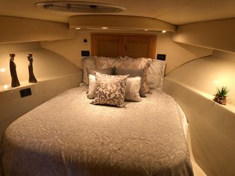 Cozy yacht cabin v-berth bedroom with cream patterned bedspread, layered decorative pillows, warm recessed lighting, a small potted plant and two sculptural accents on side shelves.