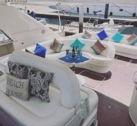Luxury yacht deck lounge with curved white seating, bright turquoise and blue throw pillows and a small table set with wine and glasses, docked at a marina.