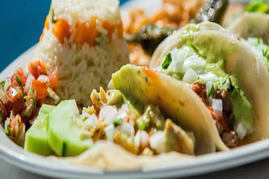Close-up of colorful Mexican tacos on a plate with diced onion and cilantro, creamy avocado sauce, pico de gallo, sliced cucumber and a molded mound of rice