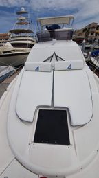 Bow of a luxury white motor yacht docked in a marina with twin cushioned sun pads and headrests trimmed in blue, a black deck hatch, neighboring boats and a partly cloudy sky.