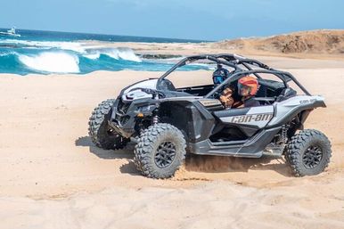 Off-road side-by-side UTV with two helmeted riders kicking up sand on a sunny coastal beach beside turquoise waves and low cliffs