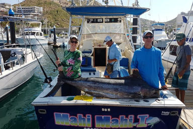 Three anglers pose with a giant marlin laid across the deck of a blue sportfishing boat at a sunny marina, with docked yachts and coastal hills in the background.
