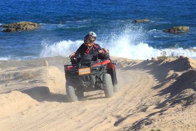 Helmeted rider on a red ATV driving along sandy coastal dunes with crashing ocean waves and rocky shoreline in the background