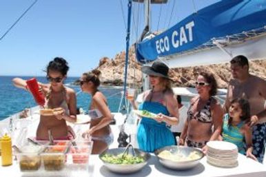 Friends and family in swimwear enjoying a buffet on a sunny sailboat, grabbing salads and condiments with clear blue sea and rocky coastline in the background.