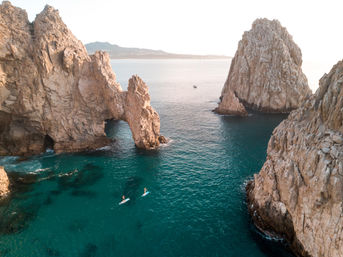 Aerial view of two stand-up paddleboarders gliding across turquoise ocean near a dramatic sunlit sea arch and towering rocky cliffs.