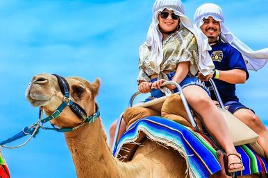 Two smiling riders wearing light head wraps and sunglasses enjoying a colorful saddle camel ride under a bright blue sky