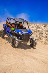 Blue four-seat off-road UTV tearing down sunlit sand dunes with two helmeted riders, kicking up a cloud of dust under a vivid blue sky.
