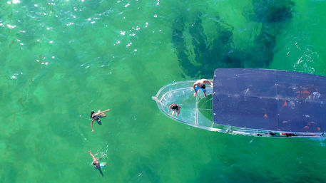 Aerial drone view of a glass-bottom boat with two people aboard and two swimmers in crystal-clear turquoise water
