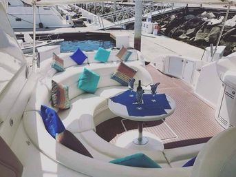 Docked luxury yacht lounge with curved white seating, teal and blue throw pillows, small round table set with a wine bottle and glasses on a teak deck, marina and rocky breakwater in the background
