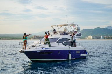 Vacationers on a sleek white-and-blue yacht enjoying calm blue coastal waters near a beachfront resort with green hills and distant mountains under a bright sky.