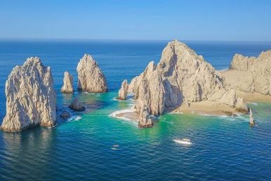Sunlit coastal rock arches and sea stacks rising from turquoise water beside a sandy cove, with a white boat and two kayaks nearby