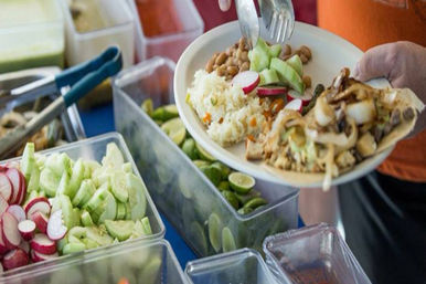Colorful taco bar plate being filled with rice, pinto beans, sautéed onions on a tortilla, sliced radishes and cucumbers, and lime wedges in plastic serving bins.