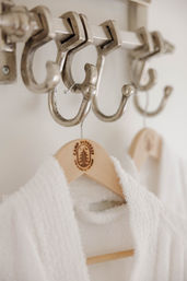 Close-up of metal wall hooks holding a wooden hanger with a tree logo and a cozy plush white bathrobe in a bright bathroom or spa.