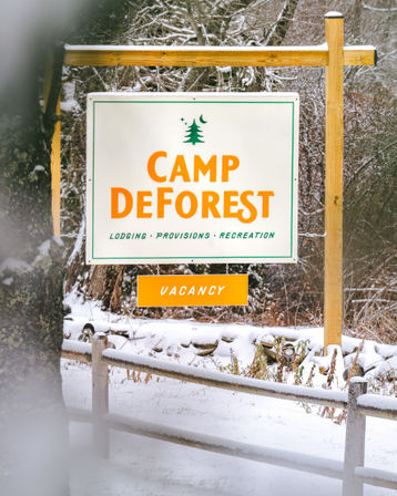 Snow-covered rustic campground sign on wooden posts advertising lodging and recreation, with a bright orange "VACANCY" tag against a snowy forest backdrop.