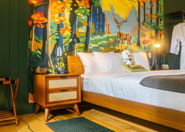 Cozy mid-century modern bedroom with wooden platform bed, white linens, rattan nightstand and lamp set against a colorful forest mural featuring a painted deer and a plush toy on the pillow.