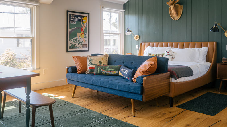 Sunlit coastal-style bedroom with a navy mid-century sofa, tan leather bed, green shiplap wall, wood floors, beach poster and decorative pillows