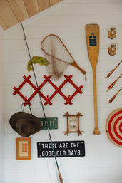 Cozy lakeside cabin wall display with a wooden oar, fishing net and rod, red folding hooks, mounted deer plaques, bullseye target, framed photo and a sign reading “These are the good old days”.