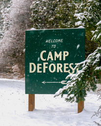 Green wooden welcome sign with an arrow, dusted in falling snow and set among snow-covered evergreen trees at a winter campground