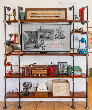 Industrial pipe-and-wood shelving styled as camp-themed display with Recreation at Camp artwork, vintage radio, lanterns, books and a plaid travel bag