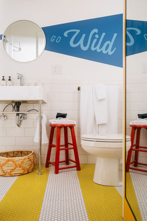 Bright, playful bathroom with white subway tile, round mirror, blue 'Go Wild' wall graphic, red wooden stool, folded towels, woven basket and yellow-and-white penny-tile floor.