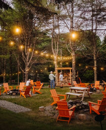 Cozy woodland outdoor lounge at dusk with orange Adirondack chairs and a picnic table around stone fire pits, warm string lights strung through pine trees and a stacked wood shelter.