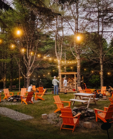 Cozy woodland outdoor lounge at dusk with orange Adirondack chairs and a picnic table around stone fire pits, warm string lights strung through pine trees and a stacked wood shelter.
