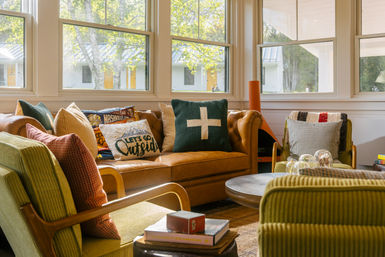 Cozy sunlit living room with a caramel leather sofa and green corduroy armchairs around a round wooden coffee table, colorful throw pillows (one reading "Let's Go Outside") and large windows overlooking a leafy yard.