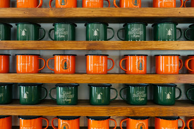 Green and orange camp-themed enamel mugs with tree graphics and "Maine" text neatly arranged on wooden display shelves