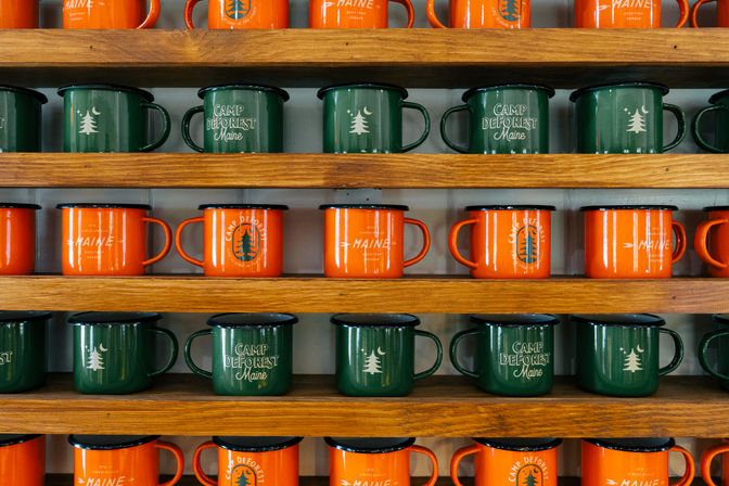 Green and orange camp-themed enamel mugs with tree graphics and "Maine" text neatly arranged on wooden display shelves