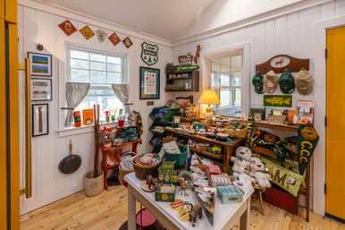 Cozy camp store interior with white beadboard walls and wooden floors, display tables and shelves overflowing with vintage camping souvenirs, hats, pennants, postcards and plush toys under warm lamp light