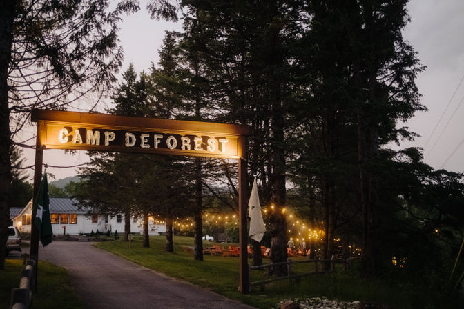 Twilight campground entrance with an illuminated wooden sign over a tree-lined driveway, cozy string lights among evergreens, rustic cabins and outdoor seating in a forest clearing.