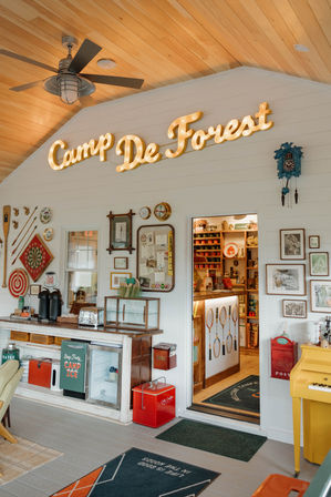Bright camp store interior with wooden ceiling fan and illuminated "Camp De Forest" sign, vintage camping decor, coolers, coffee station, framed prints and an open doorway to a cozy gift shop.