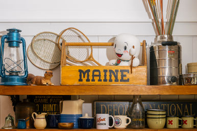 Shelf of vintage Maine souvenirs featuring a yellow wooden crate labeled MAINE with a smiling ghost toy, blue lantern, wooden tennis rackets, ceramic pitchers, mugs, and a metal canister.