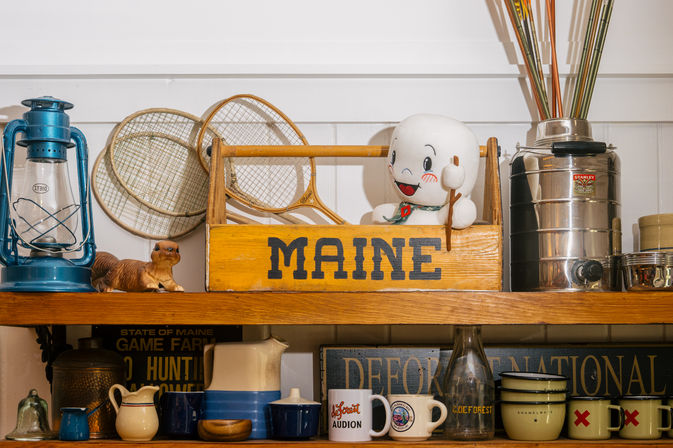 Shelf of vintage Maine souvenirs featuring a yellow wooden crate labeled MAINE with a smiling ghost toy, blue lantern, wooden tennis rackets, ceramic pitchers, mugs, and a metal canister.