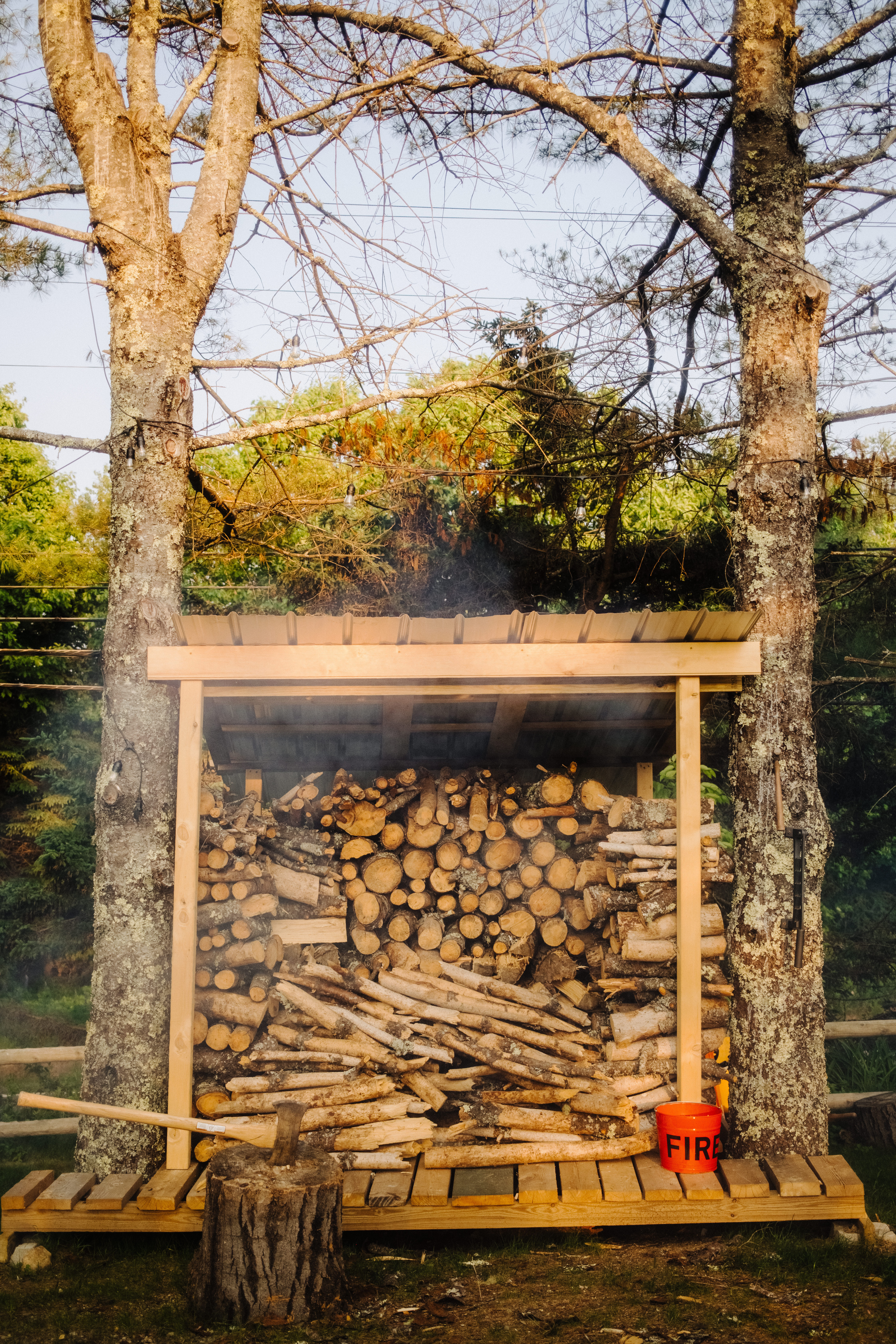 Sunlit backyard wood shelter between two trees with neatly stacked firewood and split logs, an axe in a stump and a red bucket labeled "FIRE" on the wooden platform.