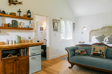 Bright cozy cottage-style bedroom with wood floors, vintage mint mini-fridge and wooden kitchenette counter, open shelves of rustic decor, teal upholstered loveseat with cabin-themed pillows, white shiplap walls and a carved wooden headboard bed by a sunny window.