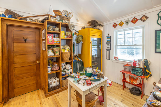 Cozy country store interior with wooden door and shelves stocked with snacks, baskets, mugs and plush toys, a bright yellow vintage refrigerator, a small white display table of local goods, sunlit window and hardwood floors.