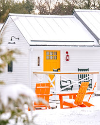 Snowy white cottage exterior with a cheerful yellow door, red lantern and bright orange Adirondack chairs on a snow-covered porch