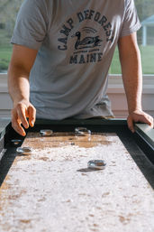 Person in a gray 'Maine' camp t‑shirt playing tabletop shuffleboard, sliding metal pucks across a sunlit, waxed wooden board dusted with shuffleboard powder.