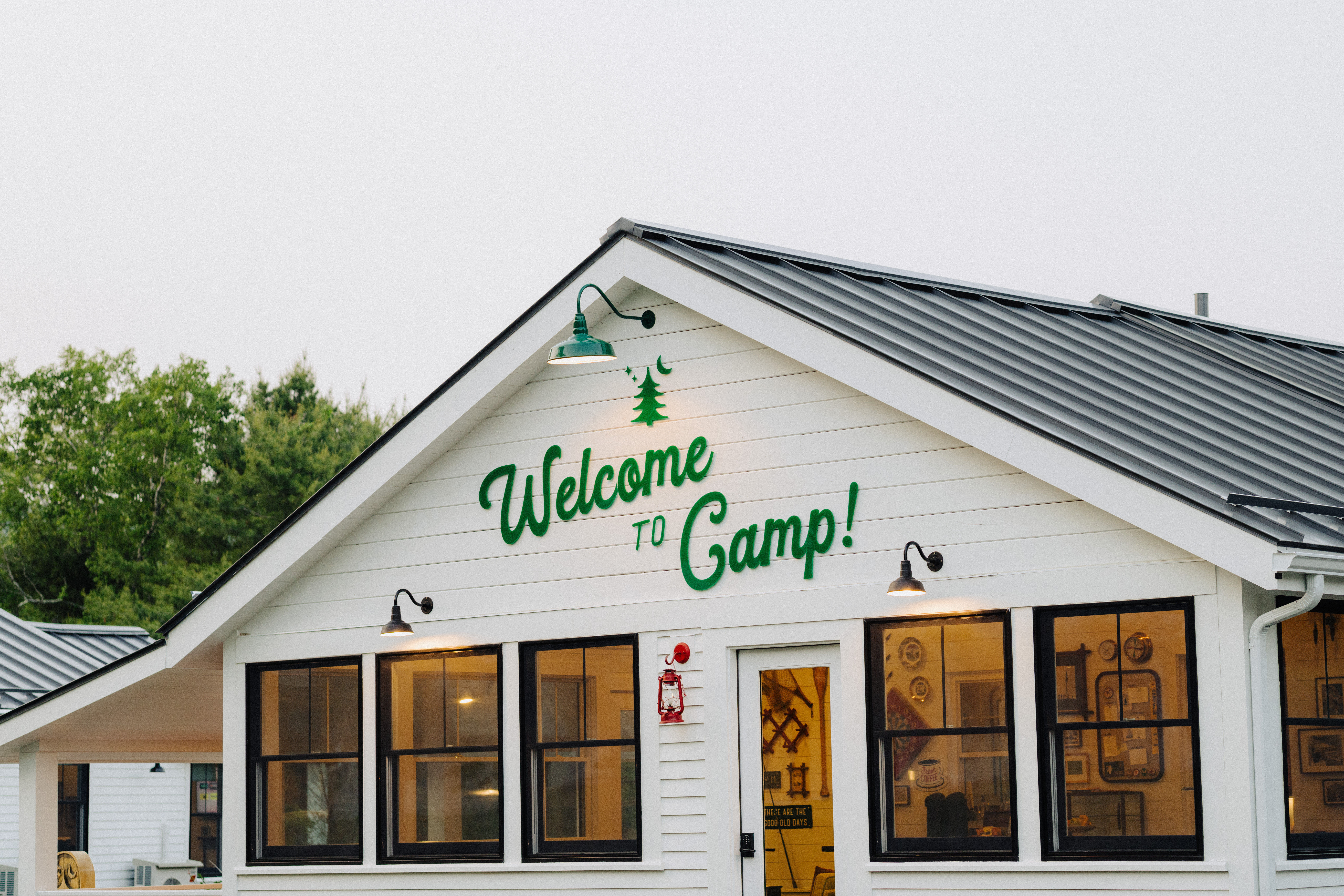 Inviting white camp lodge with green "Welcome to Camp!" lettering above the door, black-framed windows, metal roof, warm interior lights and a forest backdrop.