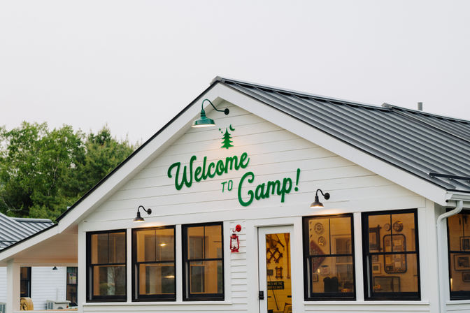 Inviting white camp lodge with green "Welcome to Camp!" lettering above the door, black-framed windows, metal roof, warm interior lights and a forest backdrop.