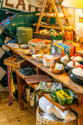 Quirky rustic gift shop display on a wooden table with vintage camping gear, pencils, patches, colorful postcards and a ceramic owl lamp.