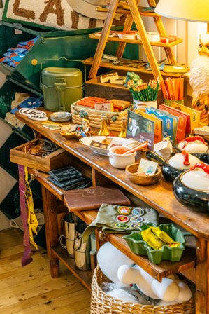 Quirky rustic gift shop display on a wooden table with vintage camping gear, pencils, patches, colorful postcards and a ceramic owl lamp.