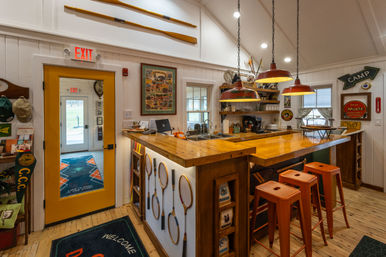 Cozy camp-lodge bar interior with glossy wooden countertop, three orange metal stools, red pendant lights, vintage wooden tennis rackets mounted on the bar, canoe paddles above, yellow exit door and rustic cabin decor