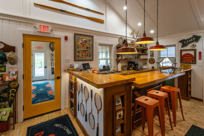 Cozy camp-lodge bar interior with glossy wooden countertop, three orange metal stools, red pendant lights, vintage wooden tennis rackets mounted on the bar, canoe paddles above, yellow exit door and rustic cabin decor