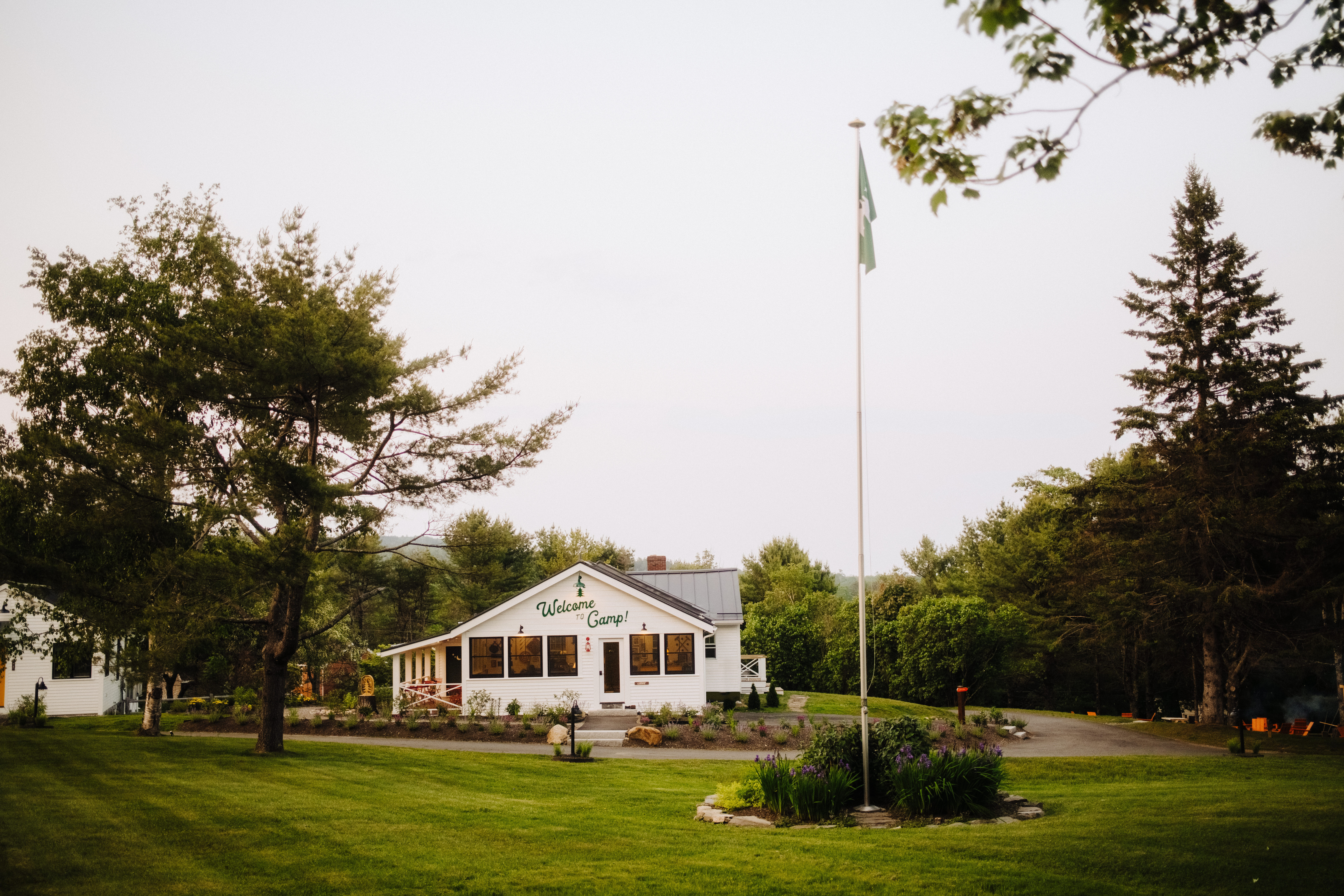 Charming white camp lodge with a 'Welcome to Camp!' sign, central flagpole, manicured green lawn and surrounding pine trees on a calm summer evening.