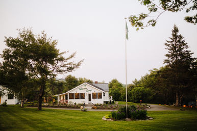 Charming white camp lodge with a 'Welcome to Camp!' sign, central flagpole, manicured green lawn and surrounding pine trees on a calm summer evening.
